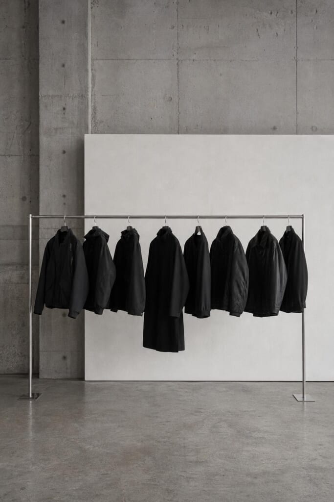 コンクリートの展示室で、白い壁の前に黒い上着が並ぶ金属のラック